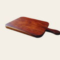 A rectangular wooden serving board with rounded corners and a single handle, shown at an angle on a plain light beige background.