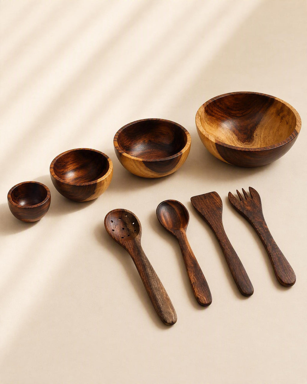A set of handcrafted wooden tableware including bowls, spoons, a spatula, and a fork, arranged on a beige background with soft diagonal shadows.