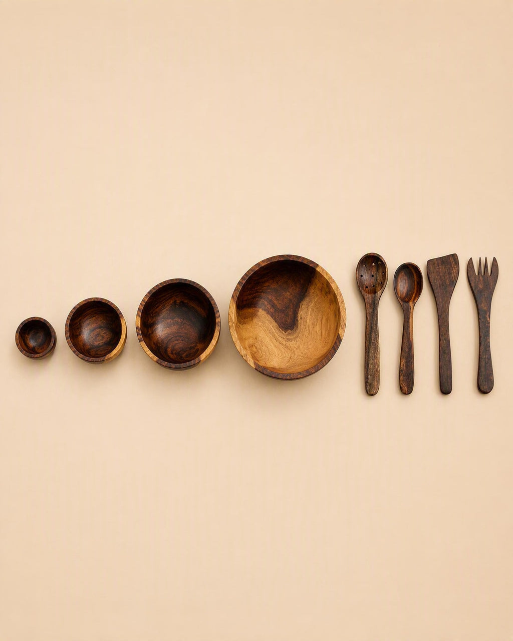 A set of dark brown wooden tableware, including bowls of decreasing size and utensils (spoon, spatula, fork), arranged in a row on a solid beige background.