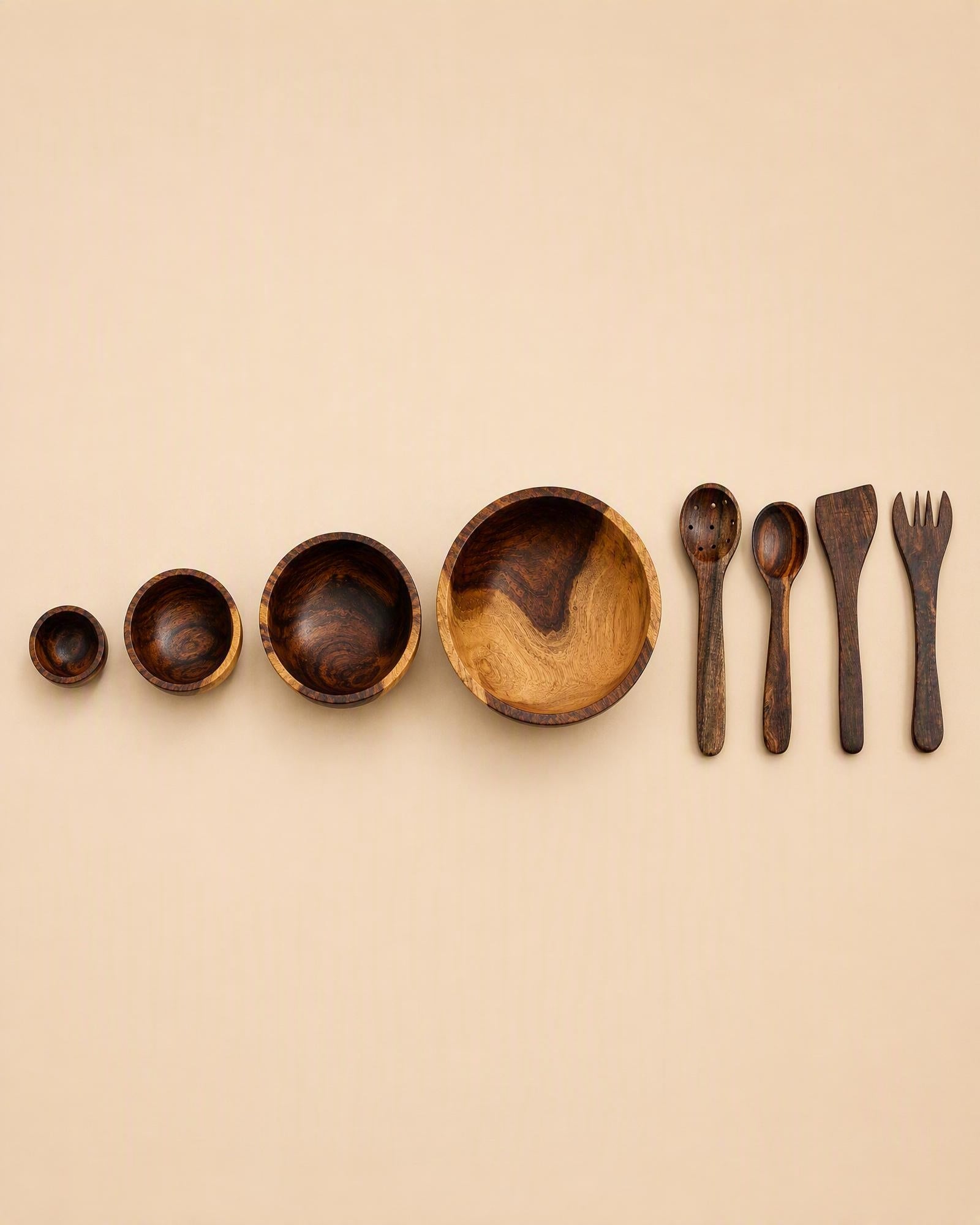 A set of dark brown wooden tableware, including bowls of decreasing size and utensils (spoon, spatula, fork), arranged in a row on a solid beige background.