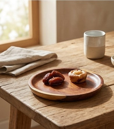 A round wooden serving plate with a natural finish, holding two dates and a small pastry, set on a rustic wooden table in a warm, sunlit room.