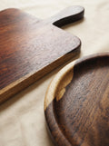 Two handcrafted wooden serving pieces, including a rectangular board with a handle and a shallow round tray, displayed on a neutral beige fabric background.