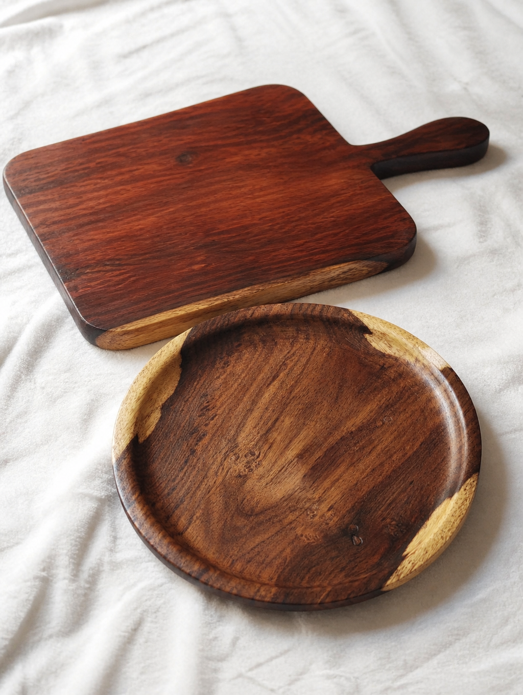 Two handcrafted wooden serving pieces on a white fabric background: a rectangular board with a handle and a round shallow tray with light wood edge accents.